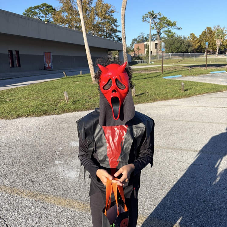 Person in costume holding bag, standing in parking lot with trees and buildings in background.