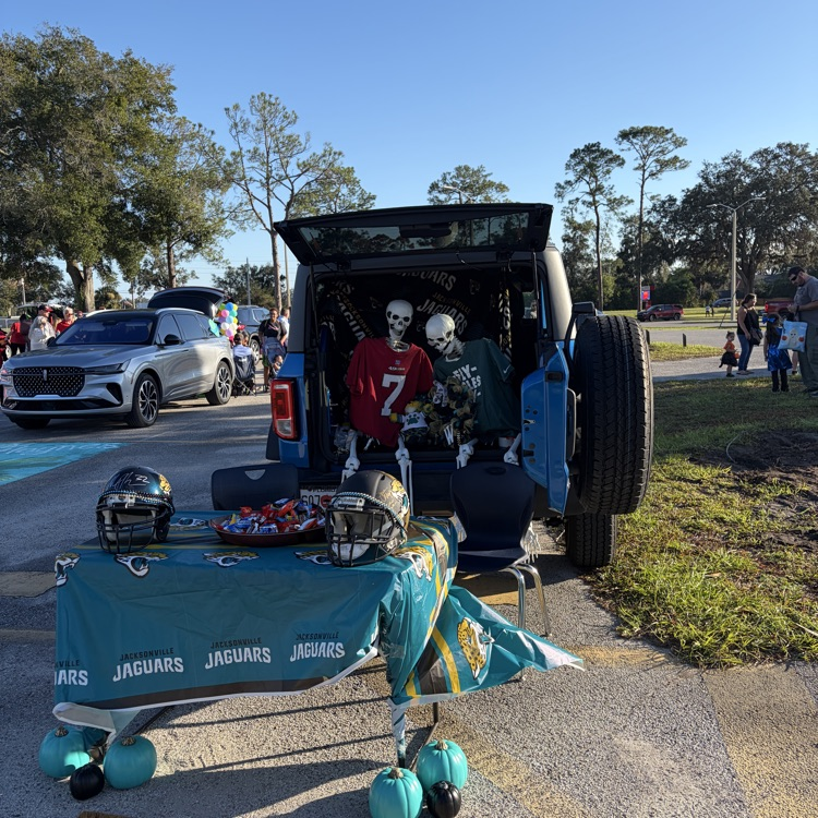 Skulls, helmets, and football jerseys in open trunk of vehicle. Table in front has balloons, pumpkins, and green tablecloth.