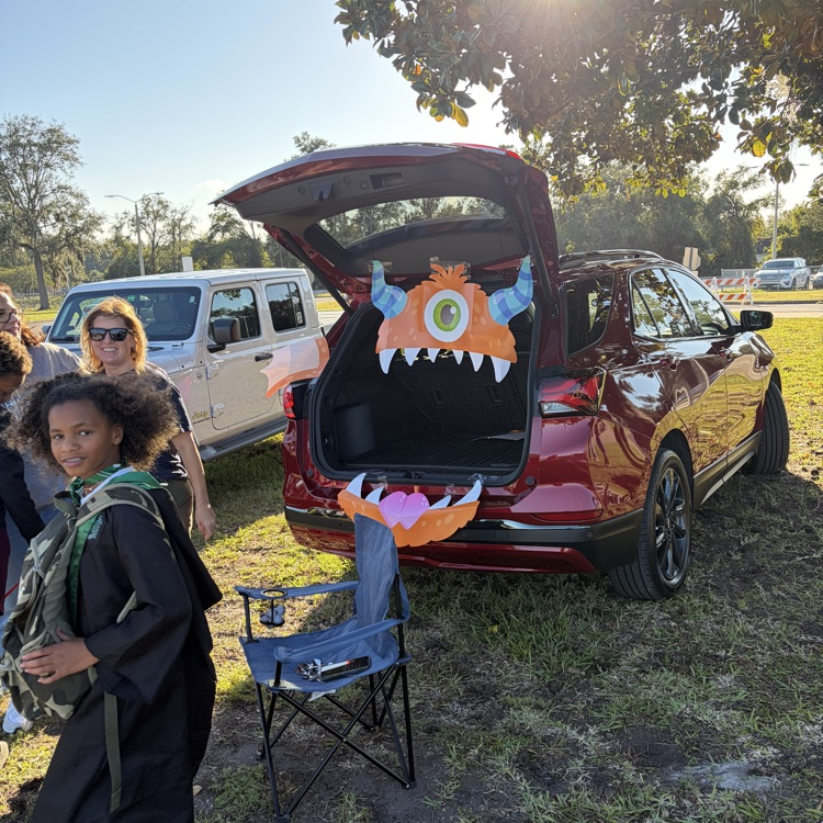 People gather near a red SUV with its trunk open, displaying a Halloween monster decoration. Nearby, a blue folding chair sits on grass.