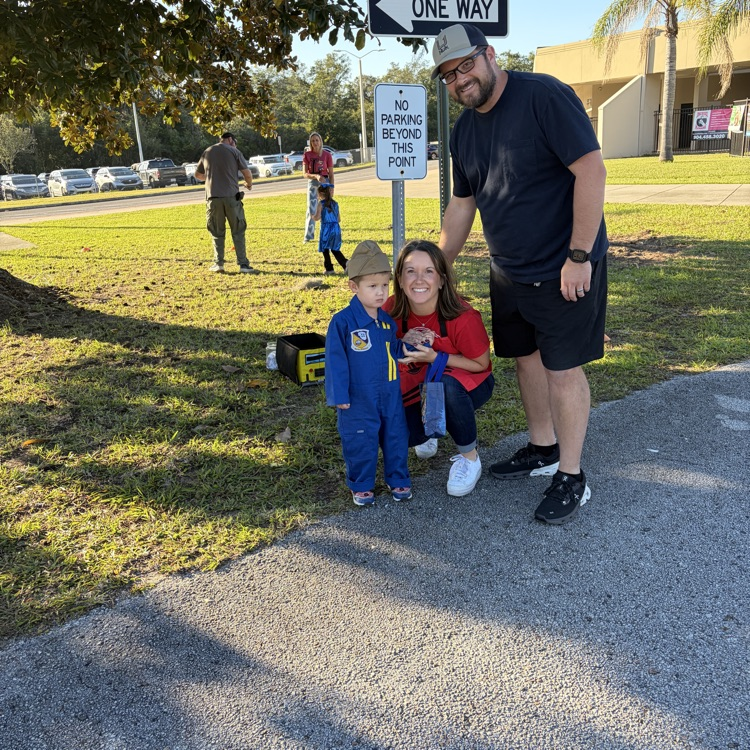 Family standing on sidewalk near one way sign with vehicles and people in background.