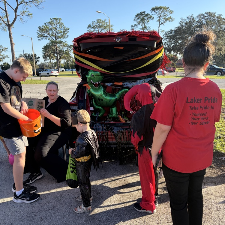 People dressed in Halloween costumes gather around a car trunk. A large dinosaur is visible in the open trunk.