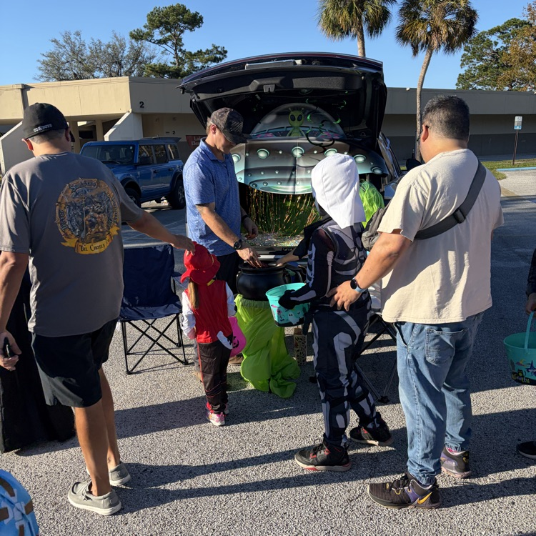 Adults and children gather near a car with the trunk open. Some people hold bags.