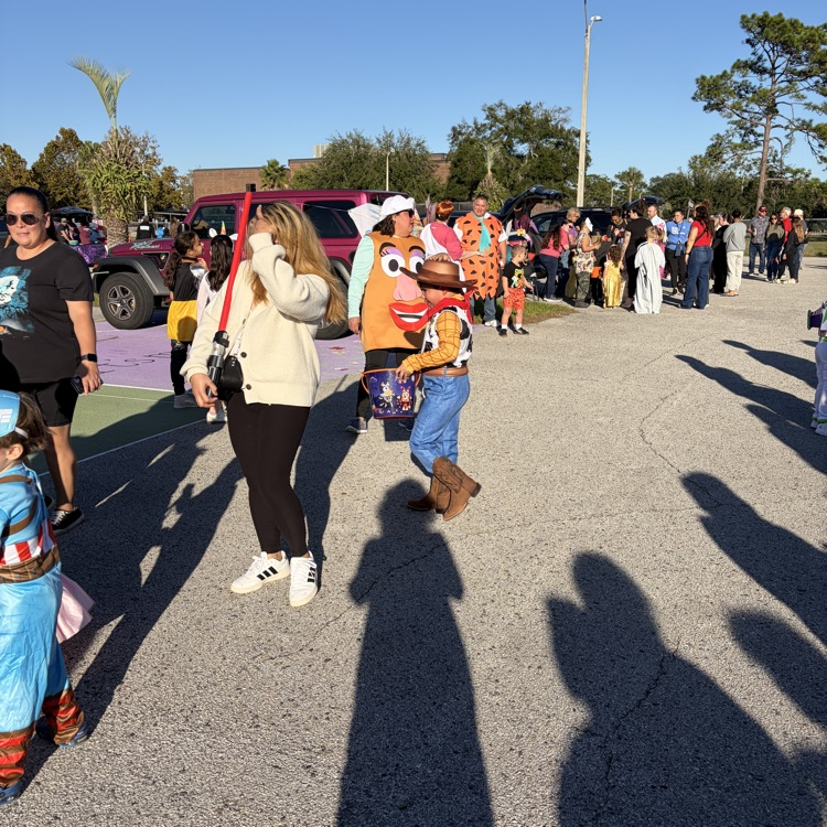 Crowds of people, some in costumes, walk along a parking lot with trees and parked cars.