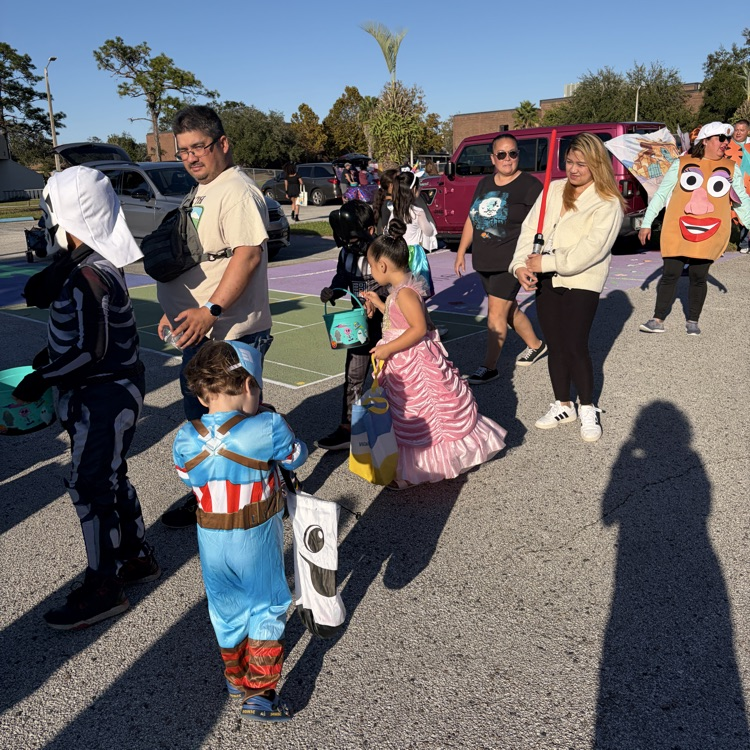 Several people, some in costumes, standing in a parking lot with cars parked and trees in background.