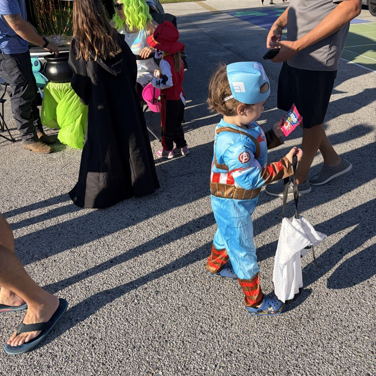 People in costumes, including a child in blue, walk on a paved path in a school setting.