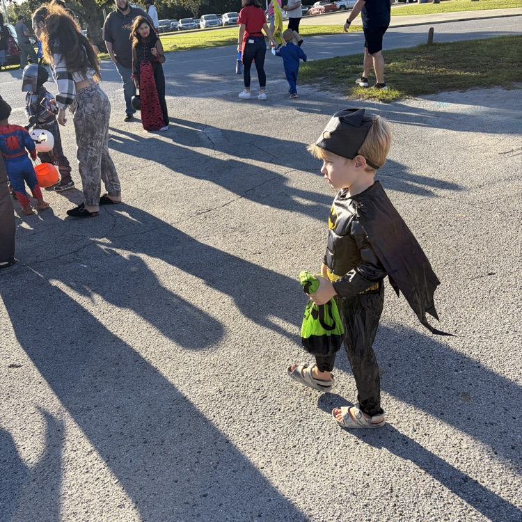 Students in costumes walk across an outdoor area. A young child in a Batman costume leads the group.