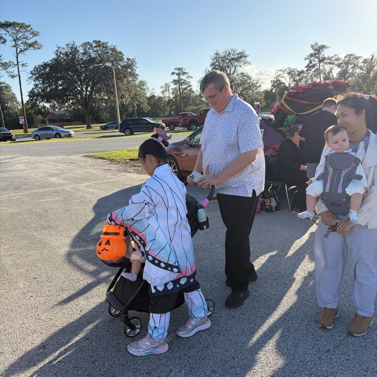People standing outdoors, a child in a pumpkin bucket, man with glasses, woman carrying baby, dark shadows, trees, parked vehicles.