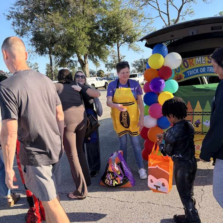 Adults and children in costumes. Person in yellow apron. Balloons. Bags. Vehicle with open trunk.