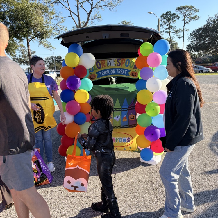 Two women and a child near a colorful balloon arch and a decorated car trunk.