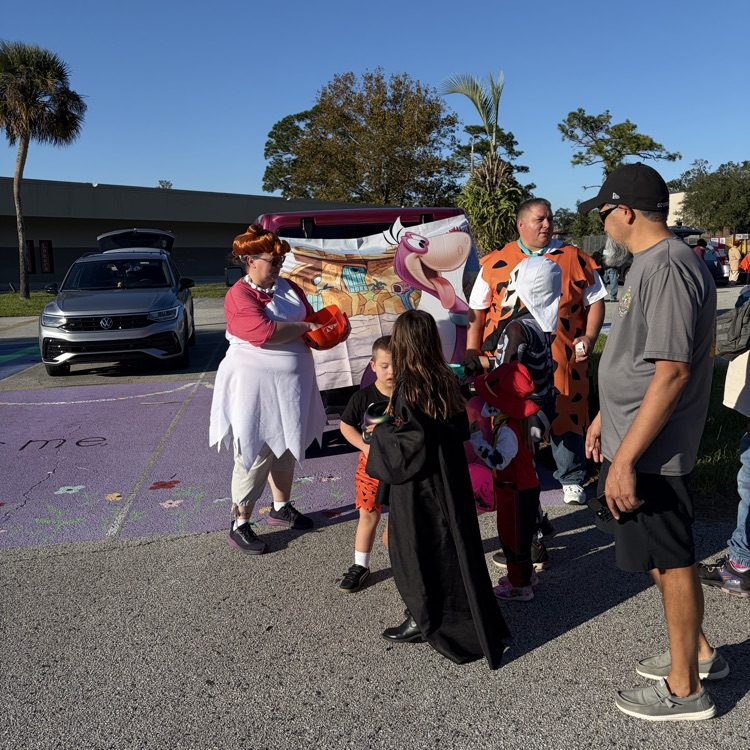 Group of adults and children dressed in costumes stand by a decorated car near a building.
