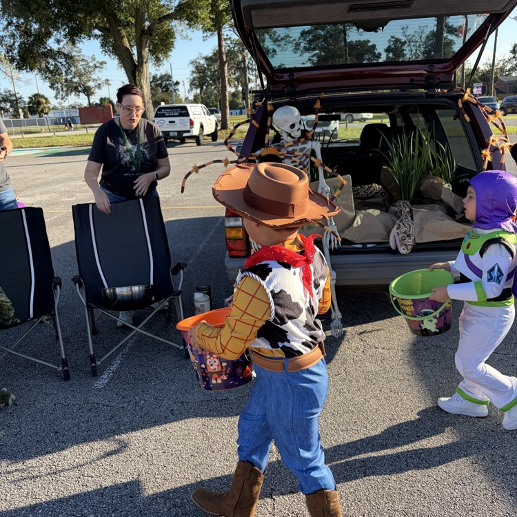 Two children in costumes with buckets, standing in parking lot, near car with open trunk, and adult woman with watch.