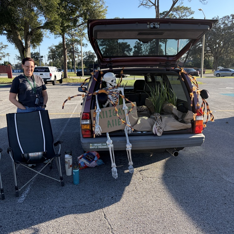 A person stands next to a chair in a parking lot. A car trunk is open with skeletons and a sign.