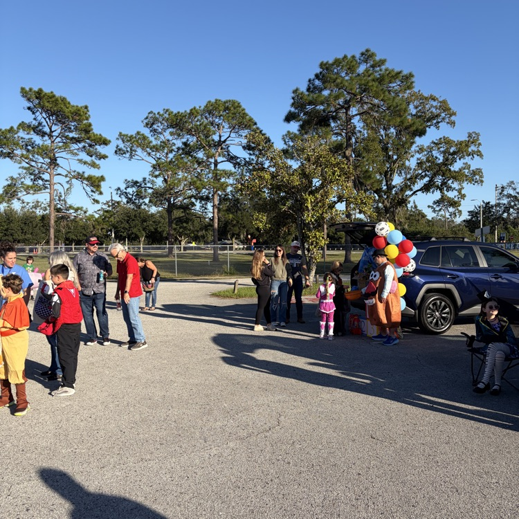 Group of people in a parking lot with a balloon bouquet, trees, and a car in the background.