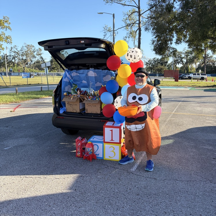 Person in costume holding a bowl, standing by car trunk filled with boxes and balloons.