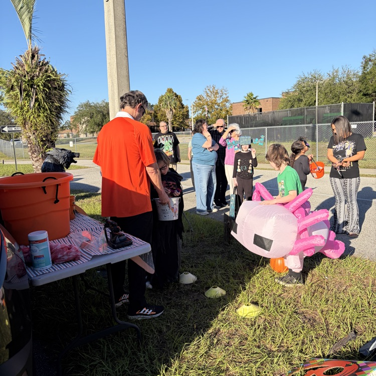 Group of children and adults outdoors. Some children holding orange pumpkins. Orange bucket on a table.