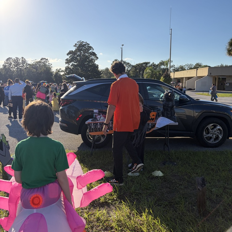 Two people in front of a parked car. One person holds a pink prop. Two people walk in background.
