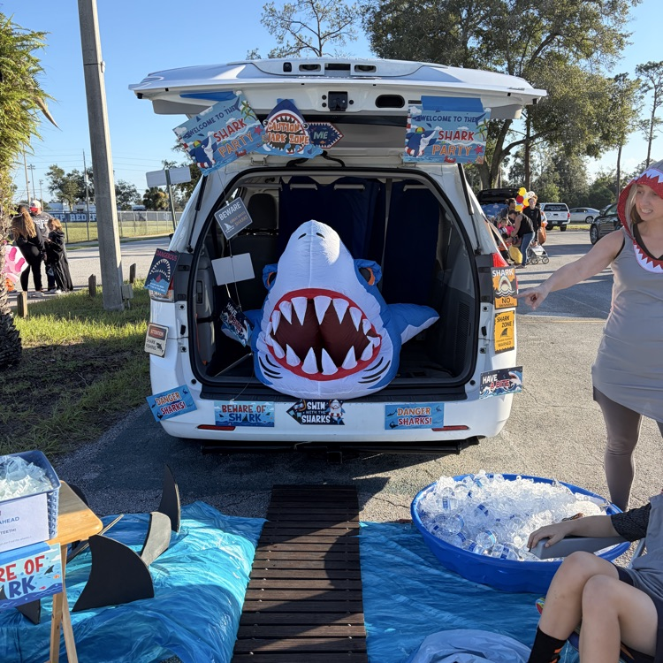 Open trunk of a car with shark inside, people in background, one person seated on ice bucket.
