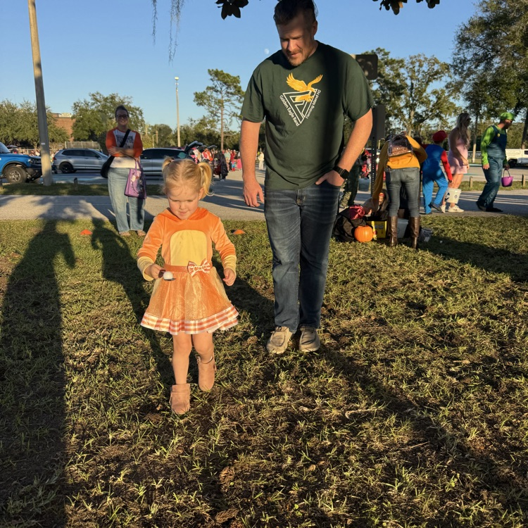 Man and girl walking on a grassy field; other people, cars, trees, and pumpkins in background.