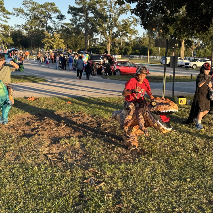 Several people dressed in dinosaur costumes walking along a road with many people walking in the background.