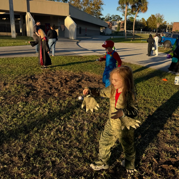 Children dressed in costumes playing on grass in front of a building; one holds an object.