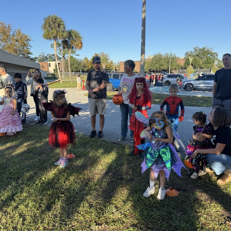 A group of children in costumes gather on a lawn. Several adults stand and sit behind them.