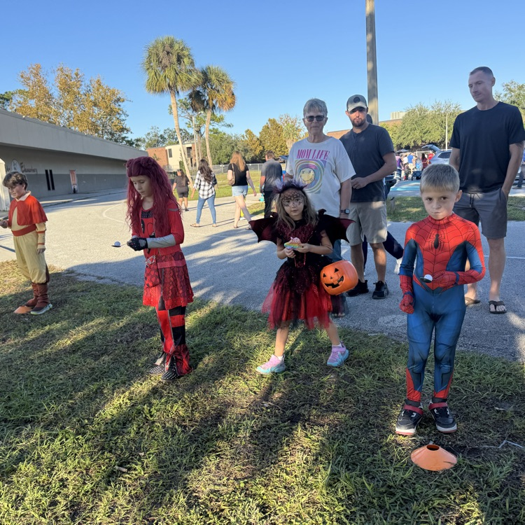 Several kids in costumes, including Spiderman and a witch, stand on grass; some hold pumpkins.