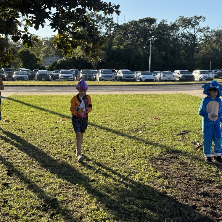 Children in costumes walk on a grassy field with cars parked on the side, and a tree provides shade.