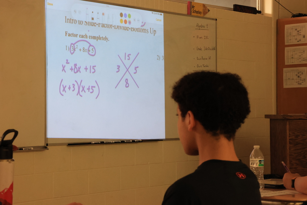 Students in a high school algebra classroom are working at their desks on problems involving rational expressions. The teacher is guiding them through a step-by-step method that includes factoring expressions, identifying common factors, and simplifying by dividing. Students are writing on paper and focusing on their work as they practice the process and check for values that cannot be used in the denominator.