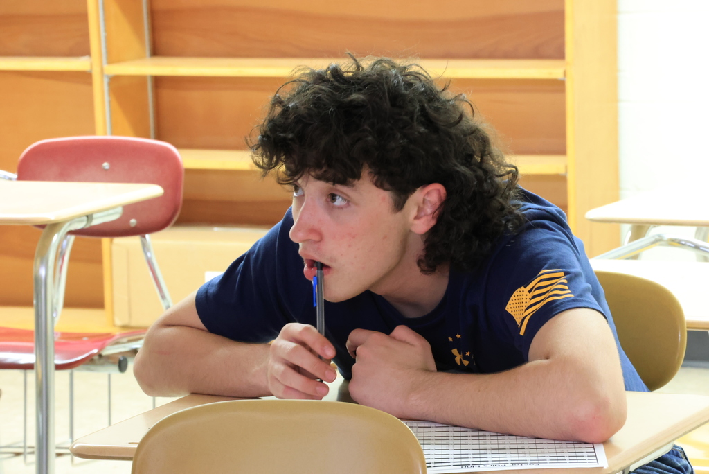 Students in a high school algebra classroom are working at their desks on problems involving rational expressions. The teacher is guiding them through a step-by-step method that includes factoring expressions, identifying common factors, and simplifying by dividing. Students are writing on paper and focusing on their work as they practice the process and check for values that cannot be used in the denominator.