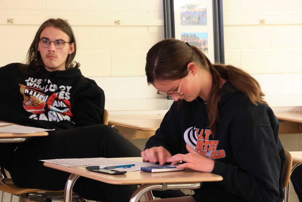 Students in a high school algebra classroom are working at their desks on problems involving rational expressions. The teacher is guiding them through a step-by-step method that includes factoring expressions, identifying common factors, and simplifying by dividing. Students are writing on paper and focusing on their work as they practice the process and check for values that cannot be used in the denominator.