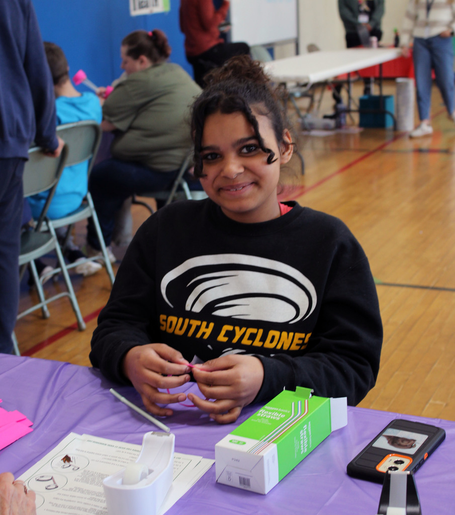 Student smiling at the camera while making a glider with paper, a straw, and tape.