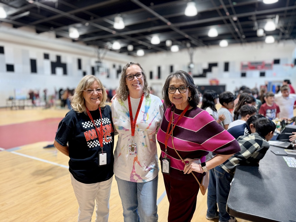 Three women stand in a gymnasium with wooden floors and overhead lighting. They wear casual clothing. A crowd of people is behind them.