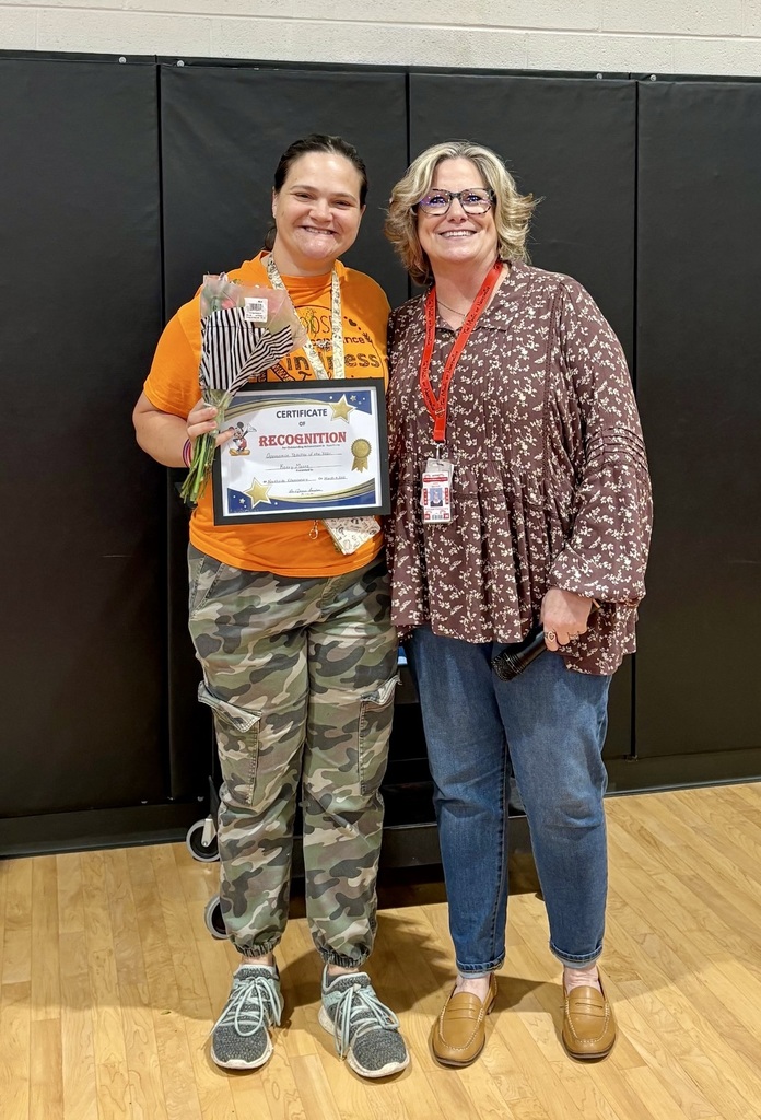 Two women stand with a trophy, one holding it and wearing a medal, both smiling. Behind them, a black wall and a wooden floor are visible.