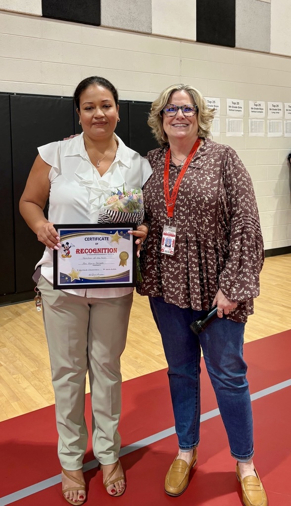 Two women stand on a red carpet in a gymnasium, one holding an award plaque, both wearing casual clothing.