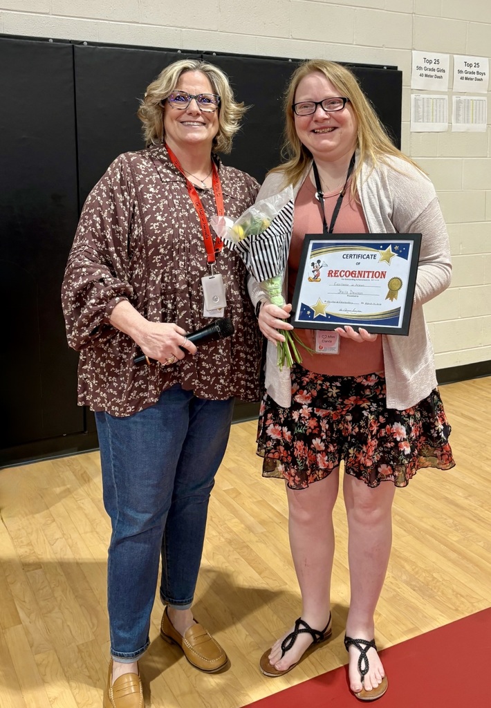 Two women stand on a wooden floor, one holding a bouquet and the other a framed certificate. Both wear ID lanyards.