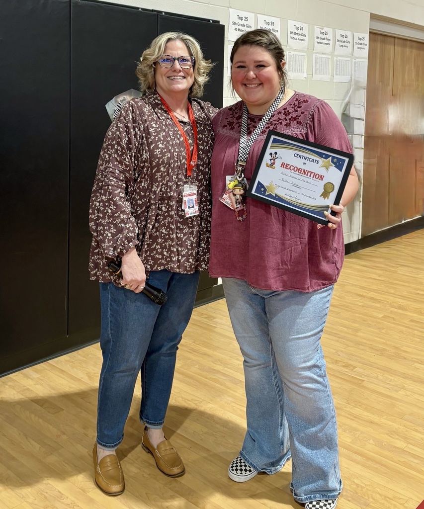 Two women stand next to each other in a hallway. The woman on the right holds a plaque.