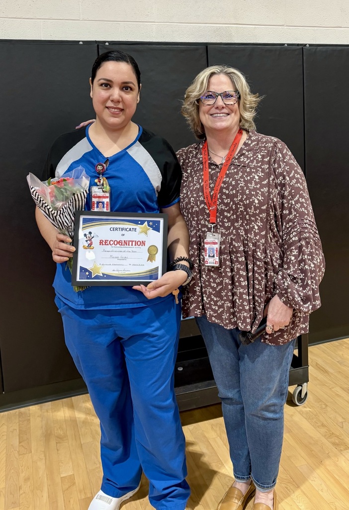 Two women stand together, one holding a framed certificate and flowers, both wearing name tags. Behind them is a black wall.