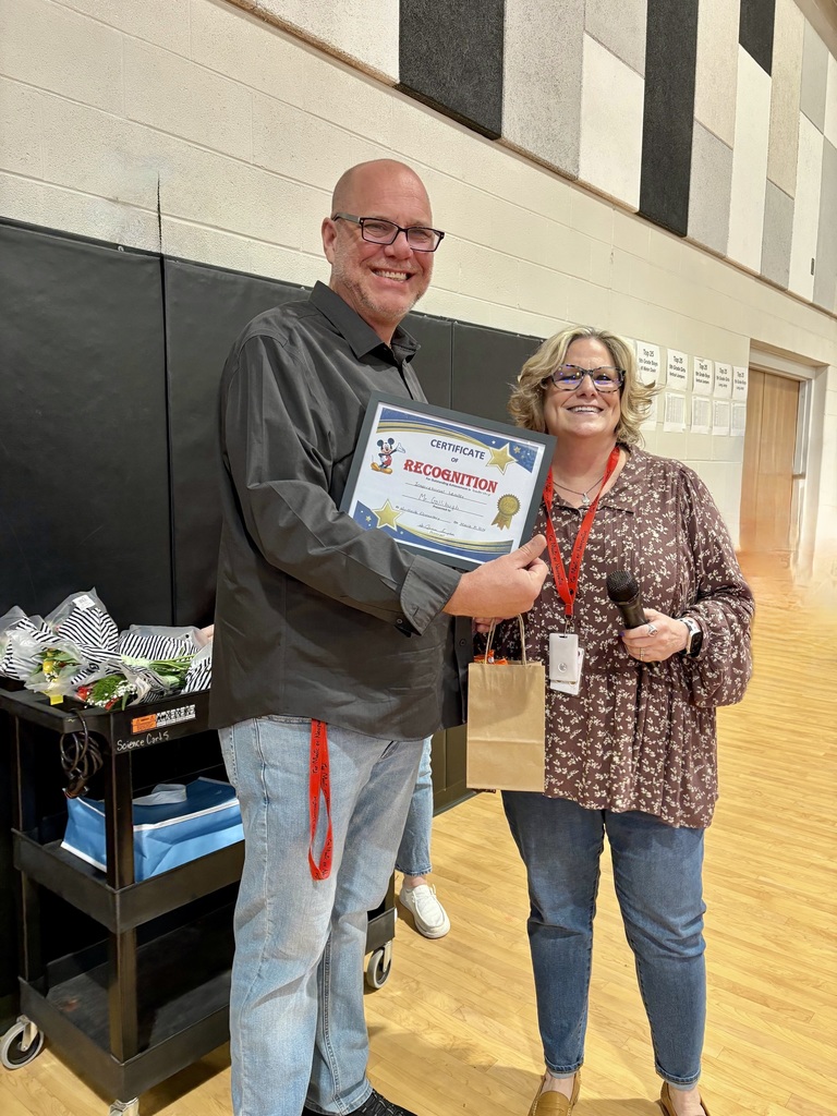 Man in glasses and woman with a paper bag holding a framed award standing in a school gym.