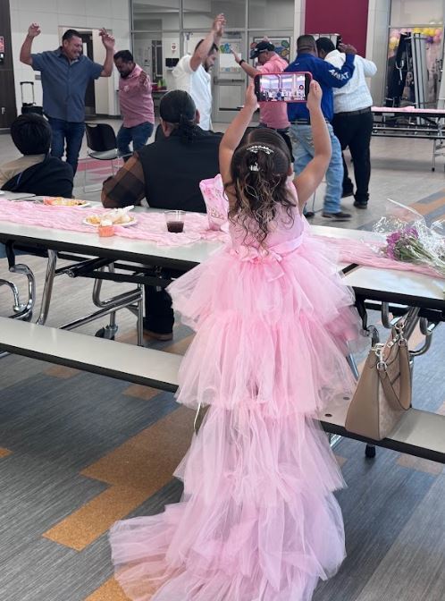 A young girl in a pink tutu dress stands in a cafeteria, surrounded by people taking photos.