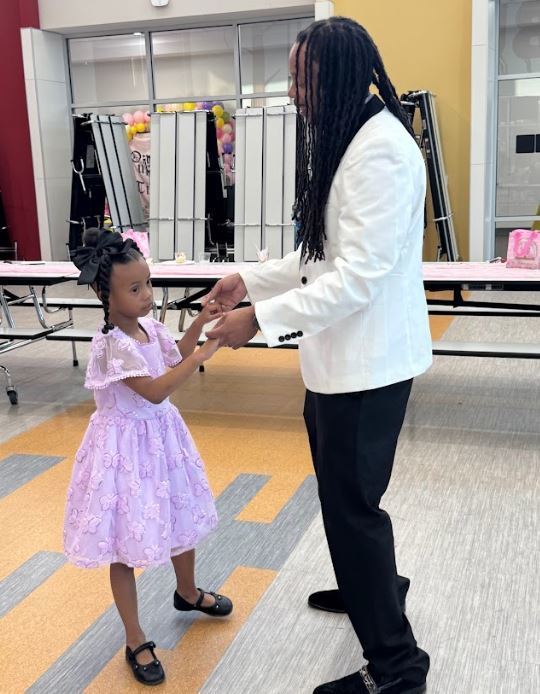 Man and girl in pink dress dance together in a room with a long table and chairs behind them.