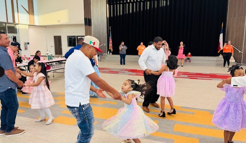 Several adults and children dancing on a floor with yellow and white stripes in an indoor hall.