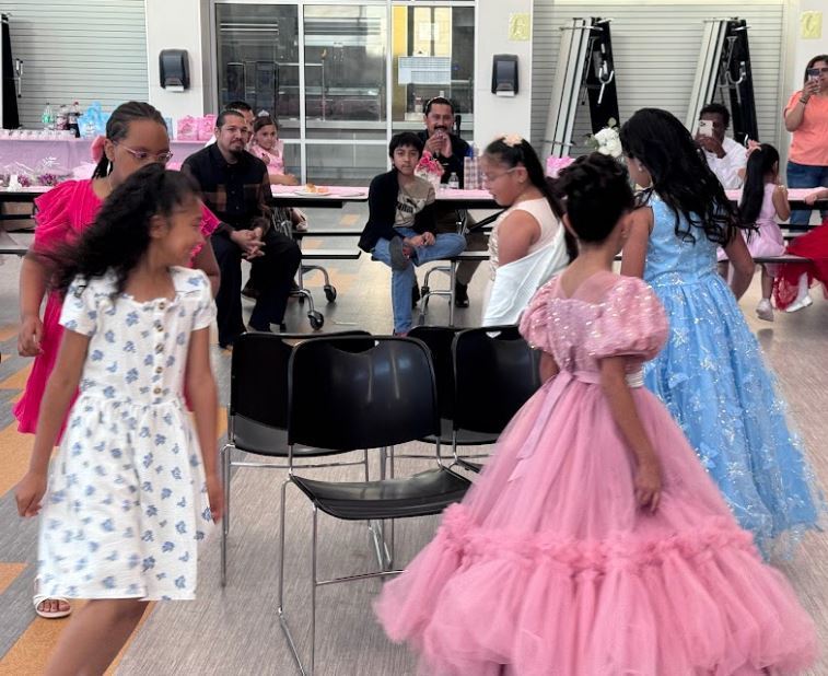 Children in colorful dresses, including pink and white, walk in a room with tables and chairs.