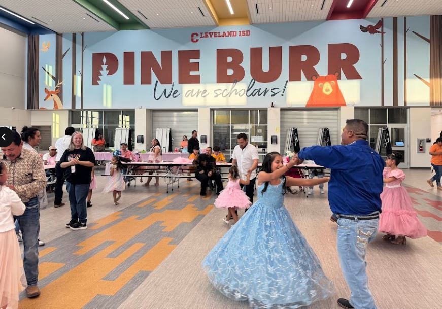 A group of people dancing in a school. One man dances with a girl in a blue dress.