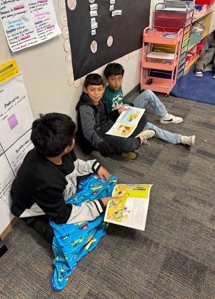 Three children sit on a carpeted floor, reading a book. Behind them, a bulletin board and a shelf with school supplies are visible.