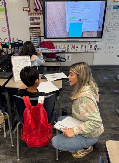A teacher crouches next to a student. Behind them, a whiteboard is visible, and a monitor shows a presentation.