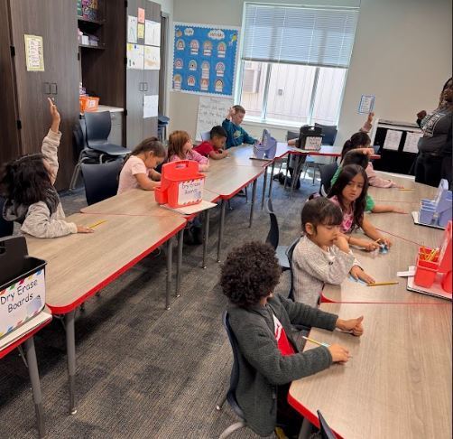 A classroom with rows of children at desks, one raising a hand, and a teacher in the background.