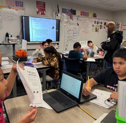 Students in a classroom, working at desks. One student holds a paper, another uses a laptop, and a teacher stands nearby.