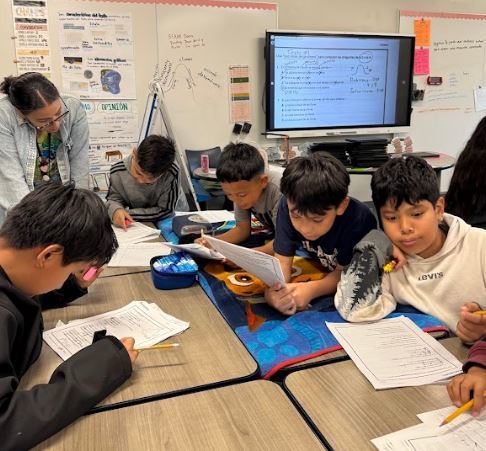 Students in a classroom work on papers at their desks with a teacher standing behind them and a monitor displaying content.