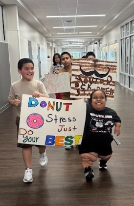A group of children holding colorful signs, one reading "DONUT Stress Do Your BEST Just." They are in a hallway.