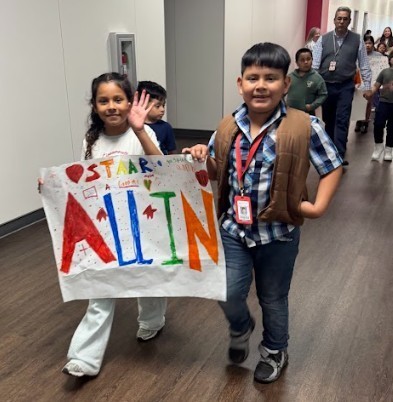 Children in a hallway, one holding a colorful banner that says "All In". Another child is smiling, and several people are in the background.